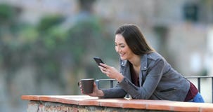 Mujer feliz que bebe y que usa el teléfono en un balcón Mujer feliz que bebe y que usa el teléfono en un balcón