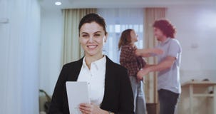 Mujer hermosa sonriente de la casa del agente que mira derecho a la cámara mientras que en fondo los pares son abrazo feliz por c metrajes Mujer hermosa sonriente de la casa del agente que mira derecho a la cámara mientras que en fondo los pares son abrazo feliz por c metrajes