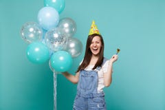 Mujer joven de risa en ropa del dril de algodón, tubo de la tenencia del sombrero del cumpleaños, celebrando con los balones de a imagen de archivo Mujer joven de risa en ropa del dril de algodón, tubo de la tenencia del sombrero del cumpleaños, celebrando con los balones de a imagen de archivo