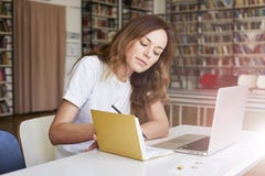 Mujer joven que trabaja en el ordenador portátil en la oficina o la biblioteca de co-trabajo, estante detrás Escritura de la empr imagen de archivo libre de regalías Mujer joven que trabaja en el ordenador portátil en la oficina o la biblioteca de co-trabajo, estante detrás Escritura de la empr imagen de archivo libre de regalías