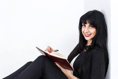 Mujer morena feliz hermosa joven que sostiene un cuaderno vestido en un traje de negocios negro que se sienta en un piso en una o imagenes de archivo Mujer morena feliz hermosa joven que sostiene un cuaderno vestido en un traje de negocios negro que se sienta en un piso en una o imagenes de archivo