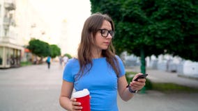 Mujer que habla en el smartphone y el café de consumición que caminan abajo de la calle Cámara lenta Mujer que habla en el smartphone y el café de consumición que caminan abajo de la calle Cámara lenta