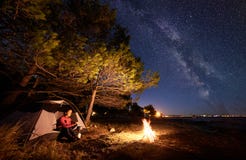 Mujer que tiene un resto en la noche que acampa cerca de la tienda turística, hoguera en orilla de mar debajo del cielo estrellad foto de archivo libre de regalías Mujer que tiene un resto en la noche que acampa cerca de la tienda turística, hoguera en orilla de mar debajo del cielo estrellad foto de archivo libre de regalías
