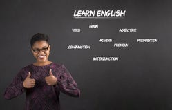 A mulher afro-americano com os polegares acima do sinal de mão aprende o inglês no fundo do quadro-negro imagem de stock A mulher afro-americano com os polegares acima do sinal de mão aprende o inglês no fundo do quadro-negro imagem de stock