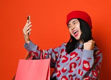 A mulher asi?tica em uma boina e em uma camiseta ? moda, com um saco de compras colorido faz um selfie trabalhador no telefone ap fotos de stock royalty free A mulher asi?tica em uma boina e em uma camiseta ? moda, com um saco de compras colorido faz um selfie trabalhador no telefone ap fotos de stock royalty free
