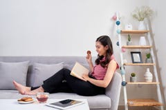 Mulher asiática bonita nova que senta-se no sofá que lê um enjo do livro fotografia de stock Mulher asiática bonita nova que senta-se no sofá que lê um enjo do livro fotografia de stock