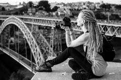 A mulher bonita nova com dreadlocks e uma câmera senta-se tomando imagens de Dom Luis que eu construo uma ponte sobre em Porto ve Fotos de Stock Royalty Free A mulher bonita nova com dreadlocks e uma câmera senta-se tomando imagens de Dom Luis que eu construo uma ponte sobre em Porto ve Fotos de Stock Royalty Free