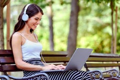 Mulher bonita nova com os fones de ouvido que sentam-se no banco no parque, usando o laptop fotos de stock Mulher bonita nova com os fones de ouvido que sentam-se no banco no parque, usando o laptop fotos de stock