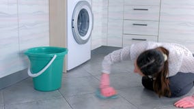 A mulher cansada em luvas de borracha cor-de-rosa lava e fricciona a mancha no assoalho da cozinha com um pano Telhas cinzentas n A mulher cansada em luvas de borracha cor-de-rosa lava e fricciona a mancha no assoalho da cozinha com um pano Telhas cinzentas n