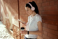 A mulher consideravelmente caucasiano dos jovens com o copo esperto do telefone e de café em suas mãos está a parede de tijolo pr fotos de stock royalty free A mulher consideravelmente caucasiano dos jovens com o copo esperto do telefone e de café em suas mãos está a parede de tijolo pr fotos de stock royalty free