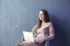 Mulher de negócios bonita que senta-se no escritório do sótão usando o portátil em joelhos Olhe e sorria Escuro - fundo azul da p fotografia de stock Mulher de negócios bonita que senta-se no escritório do sótão usando o portátil em joelhos Olhe e sorria Escuro - fundo azul da p fotografia de stock