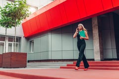 Mulher de negócios loura nova que chama o smartphone e que guarda o dobrador que anda pelo centro de negócios na cidade Communiat imagem de stock Mulher de negócios loura nova que chama o smartphone e que guarda o dobrador que anda pelo centro de negócios na cidade Communiat imagem de stock