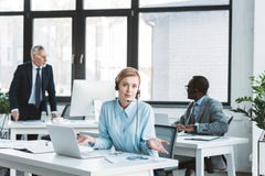 mulher de negócios nos auriculares usando o portátil e olhando a câmera quando colegas masculinos que trabalham atrás fotografia de stock royalty free mulher de negócios nos auriculares usando o portátil e olhando a câmera quando colegas masculinos que trabalham atrás fotografia de stock royalty free