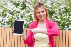 Mulher de negócios nova no estilo da elegância que senta-se no banco no parque, guardando a tela vazia da tabuleta e apontando o foto de stock Mulher de negócios nova no estilo da elegância que senta-se no banco no parque, guardando a tela vazia da tabuleta e apontando o foto de stock