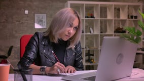 A mulher de negócios produtiva está sentando-se no local de trabalho e está escrevendo-se notas ao verificar a informação em seu filme A mulher de negócios produtiva está sentando-se no local de trabalho e está escrevendo-se notas ao verificar a informação em seu filme