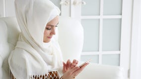 A mulher muçulmana atrativa no hijab está consultando páginas de Internet no smartphone que senta-se no sofá A mulher muçulmana atrativa no hijab está consultando páginas de Internet no smartphone que senta-se no sofá