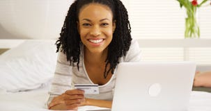 Mulher negra feliz que sorri com portátil e cartão de crédito na cama imagem de stock royalty free Mulher negra feliz que sorri com portátil e cartão de crédito na cama imagem de stock royalty free