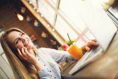 Mulher que sorri e que fala com o alguém no telefone que relaxa em um café na moda, sentando-se em uma tabela com um portátil e u Imagem de Stock Royalty Free Mulher que sorri e que fala com o alguém no telefone que relaxa em um café na moda, sentando-se em uma tabela com um portátil e u Imagem de Stock Royalty Free