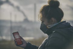 Mulher que veste uma máscara protetora contra a névoa real e que verifica a poluição do ar atual com o app esperto do telefone imagens de stock royalty free Mulher que veste uma máscara protetora contra a névoa real e que verifica a poluição do ar atual com o app esperto do telefone imagens de stock royalty free