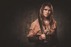 A mulher séria de viquingue com espada em um guerreiro tradicional veste-se, levantando em um fundo escuro imagens de stock A mulher séria de viquingue com espada em um guerreiro tradicional veste-se, levantando em um fundo escuro imagens de stock