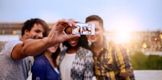 Multi-ethnic millenial group of friends taking a selfie photo with mobile phone on rooftop terrasse at sunset