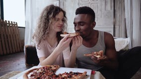 Multiracial couple have fun during the meal. Woman feed the man a slice of pizza. Male and female eating fast food.