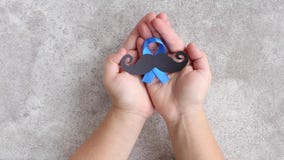 Mustache pattern with blue ribbon symbol in child`s hands. Movember concept. Prostate Cancer and men`s health awareness. Top view Mustache pattern with blue ribbon symbol in child`s hands. Movember concept. Prostate Cancer and men`s health awareness. Top view