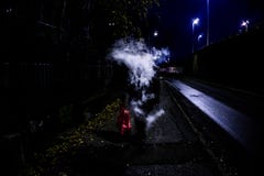 Mystisk man som utandas vaping rök som döljer dess framsida, medan gå på gatan under nattetid royaltyfri bild Mystisk man som utandas vaping rök som döljer dess framsida, medan gå på gatan under nattetid royaltyfri bild