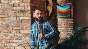 Mâle barbu beau de hippie dans une chemise bleue et des jeans d'ouatine avec le sac à dos se penchant contre un mur de briques à photo libre de droits Mâle barbu beau de hippie dans une chemise bleue et des jeans d'ouatine avec le sac à dos se penchant contre un mur de briques à photo libre de droits