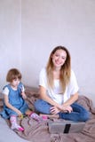 Mãe de sorriso nova que senta-se na cama com a filha pequena perto do portátil foto de stock royalty free Mãe de sorriso nova que senta-se na cama com a filha pequena perto do portátil foto de stock royalty free