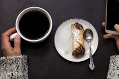 A mão caucasiano da mulher guarda o copo de café e o smartphone com o cannoli italiano na placa no fundo preto fotografia de stock A mão caucasiano da mulher guarda o copo de café e o smartphone com o cannoli italiano na placa no fundo preto fotografia de stock