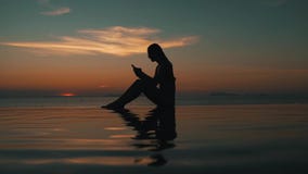 Mädchen benutzt einen Smartphone bei Sonnenuntergang durch den Ozean stock video Mädchen benutzt einen Smartphone bei Sonnenuntergang durch den Ozean stock video