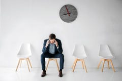 Müder und erschöpfter junger Mann sitzen auf weißem Stuhl auf Raum allein Er hält Hände auf Kopf Kerl hat Kopfschmerzen Er leidet lizenzfreies stockbild Müder und erschöpfter junger Mann sitzen auf weißem Stuhl auf Raum allein Er hält Hände auf Kopf Kerl hat Kopfschmerzen Er leidet lizenzfreies stockbild