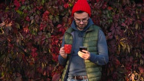 Mężczyzna w czerwonej nakrętce z filiżanką w jego rękach pije outdoors, używa smartphone na ciepłym jesień dniu i herbaty lub kaw zdjęcie wideo Mężczyzna w czerwonej nakrętce z filiżanką w jego rękach pije outdoors, używa smartphone na ciepłym jesień dniu i herbaty lub kaw zdjęcie wideo