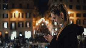 Młoda atrakcyjna kobiety pozycja w centrum miasta przy wieczór i używa smartphone Tłum i światła na tle fotografia royalty free Młoda atrakcyjna kobiety pozycja w centrum miasta przy wieczór i używa smartphone Tłum i światła na tle fotografia royalty free