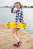 Młoda rozochocona dziewczyna w modnisia stroju mienia żółtym longboard w jego ręce i odprowadzenie na drewnianym molu zdjęcie royalty free Młoda rozochocona dziewczyna w modnisia stroju mienia żółtym longboard w jego ręce i odprowadzenie na drewnianym molu zdjęcie royalty free