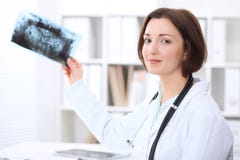 Młodej brunetki dentysty żeński obsiadanie przy stołem i egzamininuje stomatologicznego promieniowanie rentgenowskie fotografia stock Młodej brunetki dentysty żeński obsiadanie przy stołem i egzamininuje stomatologicznego promieniowanie rentgenowskie fotografia stock