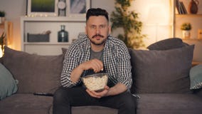 Młody brodaty mężczyzna ogląda TV i je popkorn siedzi na leżance w domu zbiory Młody brodaty mężczyzna ogląda TV i je popkorn siedzi na leżance w domu zbiory