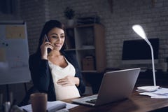 Młody kobieta w ciąży opowiada na telefonie przy biurkiem w ciemnym środowisku Kobieta w ciąży pracuje na laptopie obrazy stock Młody kobieta w ciąży opowiada na telefonie przy biurkiem w ciemnym środowisku Kobieta w ciąży pracuje na laptopie obrazy stock