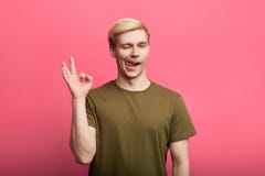 Młody życzliwy powabny facet mruga podczas gdy pokazywać OK gest, odosobnionego na różowym tle fotografia stock Młody życzliwy powabny facet mruga podczas gdy pokazywać OK gest, odosobnionego na różowym tle fotografia stock
