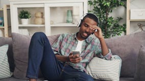 Netter Afroamerikanerkerl ist, hörend singend und Musik in den Kopfhörern, die auf Couch in der modernen Wohnung sich entspannen stock video footage Netter Afroamerikanerkerl ist, hörend singend und Musik in den Kopfhörern, die auf Couch in der modernen Wohnung sich entspannen stock video footage