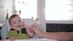 Nettes lächelndes Babyessen, zu Hause sitzend am Kindersitz Bemuttern Sie die Fütterung ihres entzückenden Jährigen mit einem Löf stock footage Nettes lächelndes Babyessen, zu Hause sitzend am Kindersitz Bemuttern Sie die Fütterung ihres entzückenden Jährigen mit einem Löf stock footage