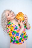 Nettes molliges Mädchen mit dem weißen gelockten Haar in einer Sommerjacke mit Blumen hält ein süßes Gebäckbrötchen in Form eines stockfotografie Nettes molliges Mädchen mit dem weißen gelockten Haar in einer Sommerjacke mit Blumen hält ein süßes Gebäckbrötchen in Form eines stockfotografie