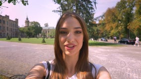 Nice young woman is talking in videochat in park in daytime, holding camera, communication concept