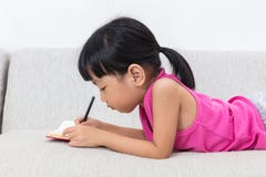 Niña china asiática que pone en el libro de la escritura del sofá Fotografía de archivo Niña china asiática que pone en el libro de la escritura del sofá Fotografía de archivo