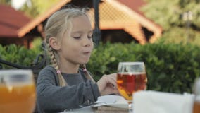 Niña con dos trenzas que se sientan en la tabla en el café al aire libre almacen de metraje de vídeo Niña con dos trenzas que se sientan en la tabla en el café al aire libre almacen de metraje de vídeo