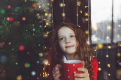 Niña linda que se sienta con una taza de cacao caliente por la ventana Imagen de archivo Niña linda que se sienta con una taza de cacao caliente por la ventana Imagen de archivo