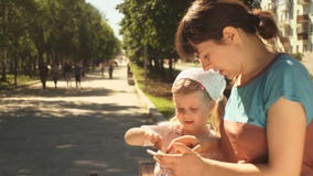 Niña y su madre que gozan de un teléfono que se sienta en un banco en el parque Niña y su madre que gozan de un teléfono que se sienta en un banco en el parque