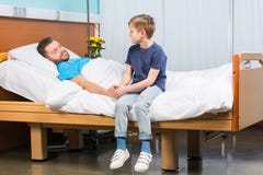 Niño pequeño que se sienta en cama de hospital y que mira al padre enfermo fotografía de archivo Niño pequeño que se sienta en cama de hospital y que mira al padre enfermo fotografía de archivo