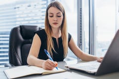 Notas de trabalho concentradas da escrita da mulher de negócios no caderno que senta-se na mesa no escritório fotos de stock Notas de trabalho concentradas da escrita da mulher de negócios no caderno que senta-se na mesa no escritório fotos de stock